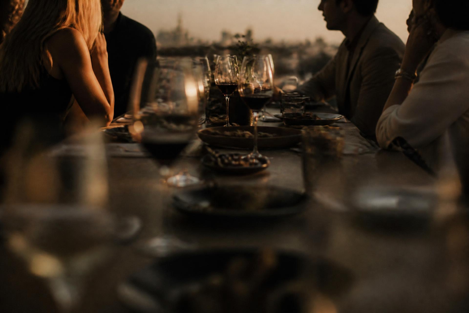 Mesa longa ao final da tarde, conversas e copos em primeiro plano desfocado